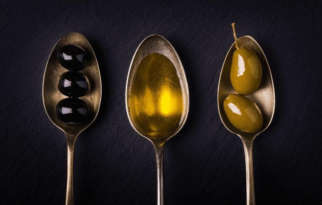 Tres cucharas antiguas sobre pizarra oscura con aceitunas negras, aceite y aceitunas verdes
