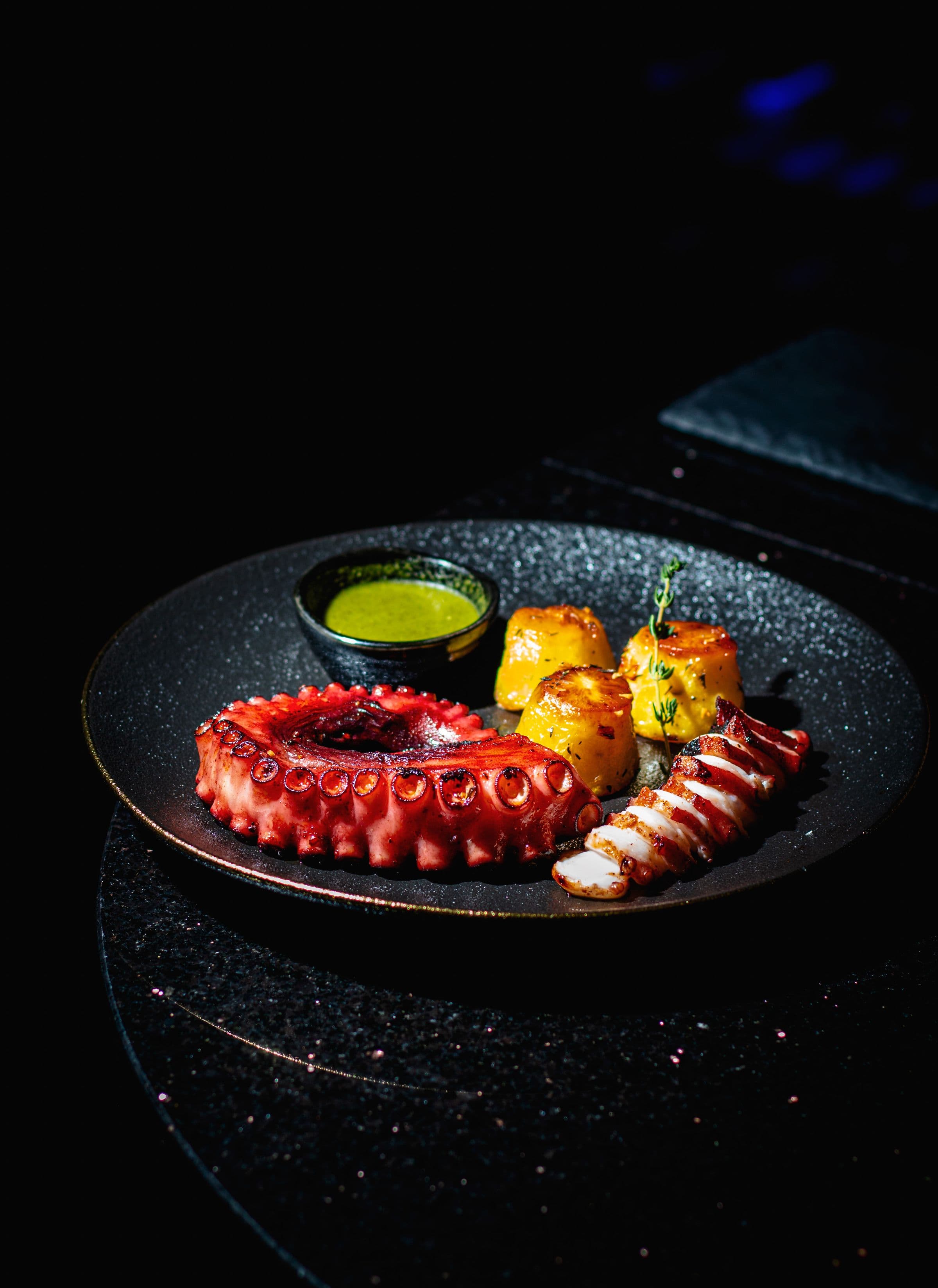 Pulpo a la brasa emplatado sobre fondo negro, con salsa verde y guarnición