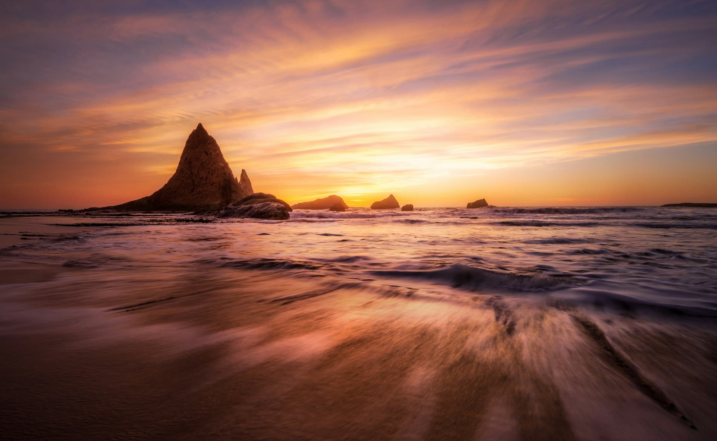 Roquedos y agujas de piedra en la orilla, olas rompiendo bajo un cielo de atardecer dorado