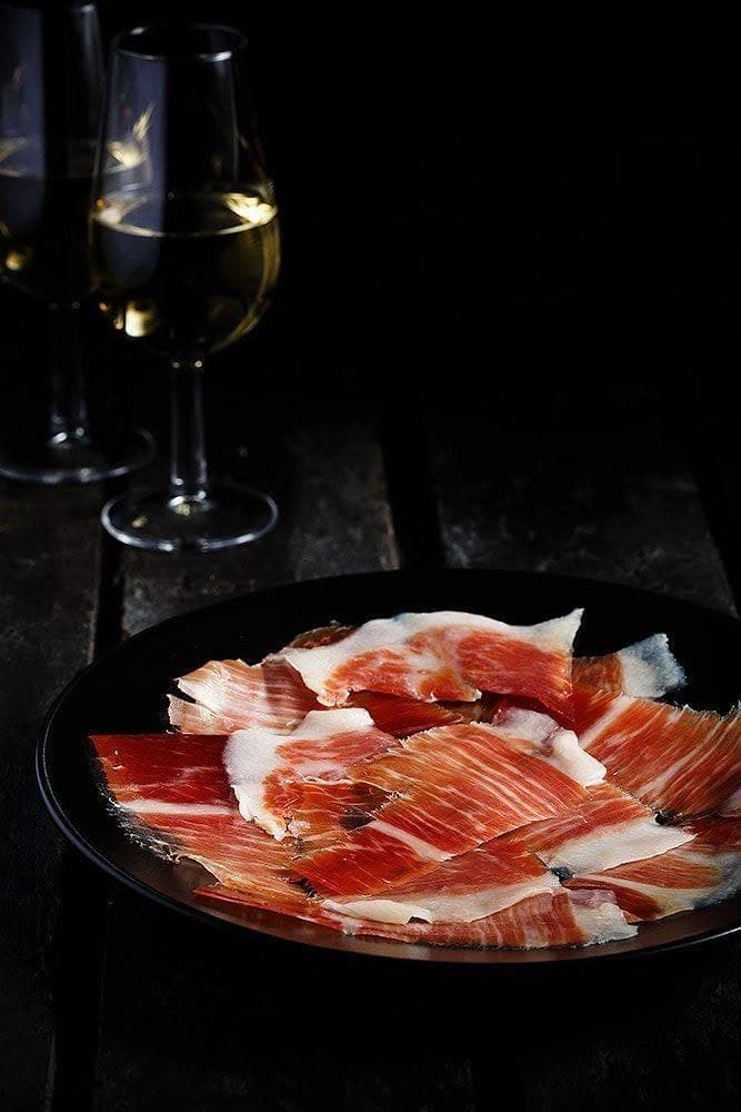 Jamón ibérico loncheado sobre plato oscuro, dos copas de vino blanco desenfocadas al fondo
